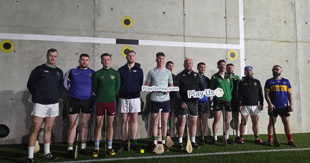 A group of adult players in different club kits standing in a line against a wall, holding Play Us signs